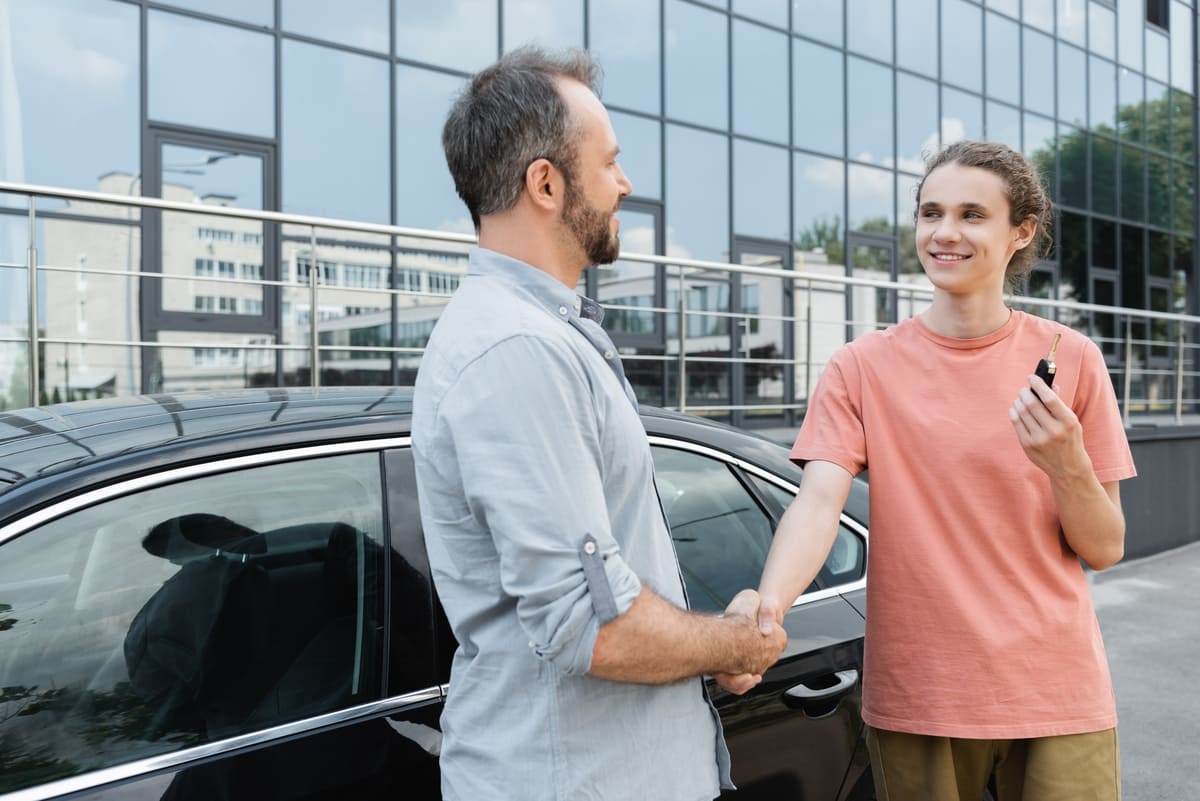 Liikluskindlustuse automaatsest üleminekust - kaks heledates riietes inimest suruvad musta auto juures rõõmsalt kätt
